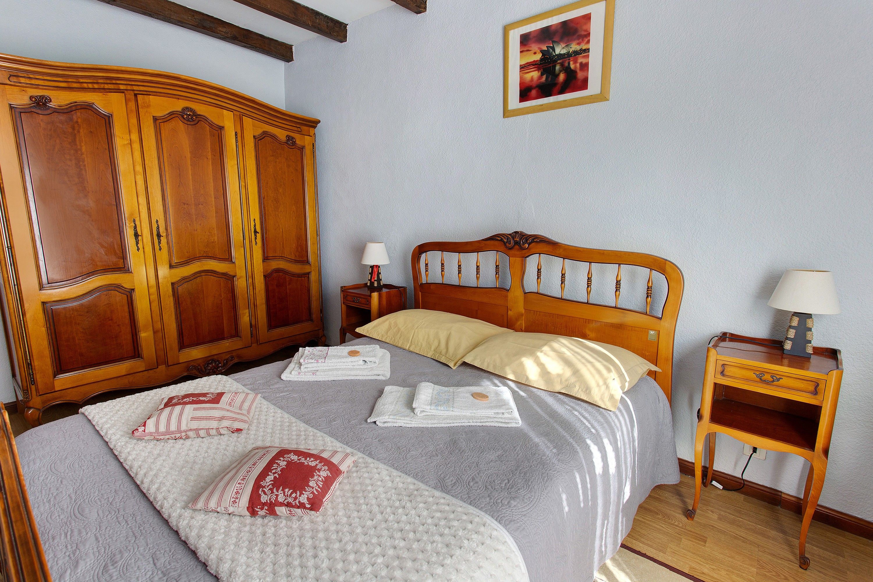 Een hotelkamer met een grote houten garderobe, een bed met quilt en kussens, een nachtkastje met een lamp en een lijst met een foto aan de muur.