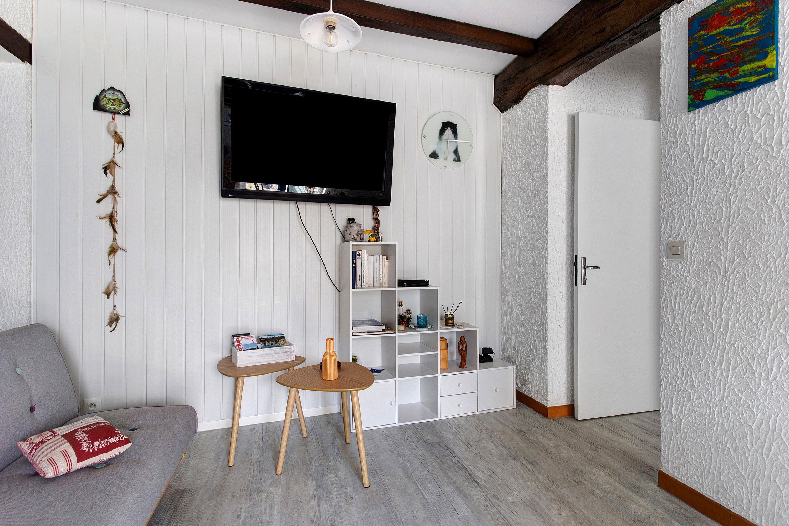 Una sala de estar con un televisor de pantalla plana montado en la pared, una estantería, una mesita con un jarrón y un sofá gris con un cojín decorativo.