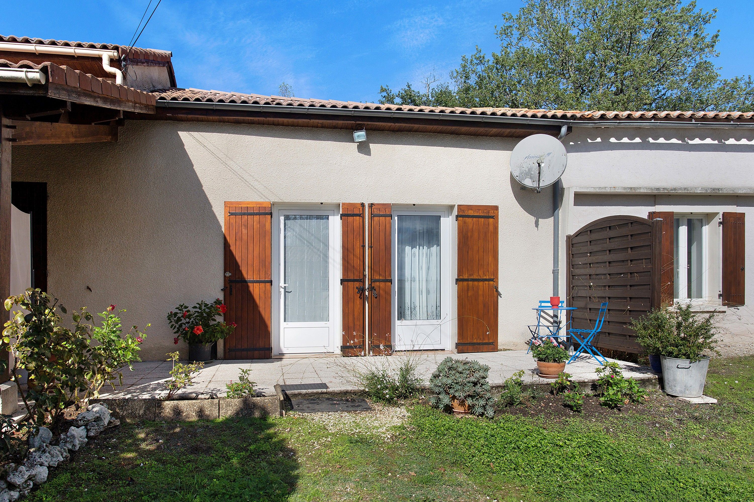 Una casa con techo de tejas, antena parabólica, puerta de madera con contraventanas y un pequeño jardín con plantas en macetas.