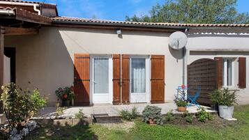 Una casa con techo de tejas, antena parabólica, puerta de madera con contraventanas y un pequeño jardín con plantas en macetas.