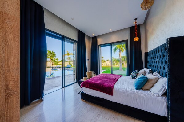 A modern bedroom with a large bed, dark curtains, and a view of a pool and palm trees. - Spacious 5-bedroom villa in peaceful Marrakech-Safi with AC (Marrakech-Safi)