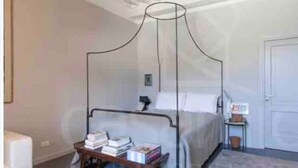 Een slaapkamer met een hemelbed, een nachtkastje met boeken, een nachtkastje en een deur.