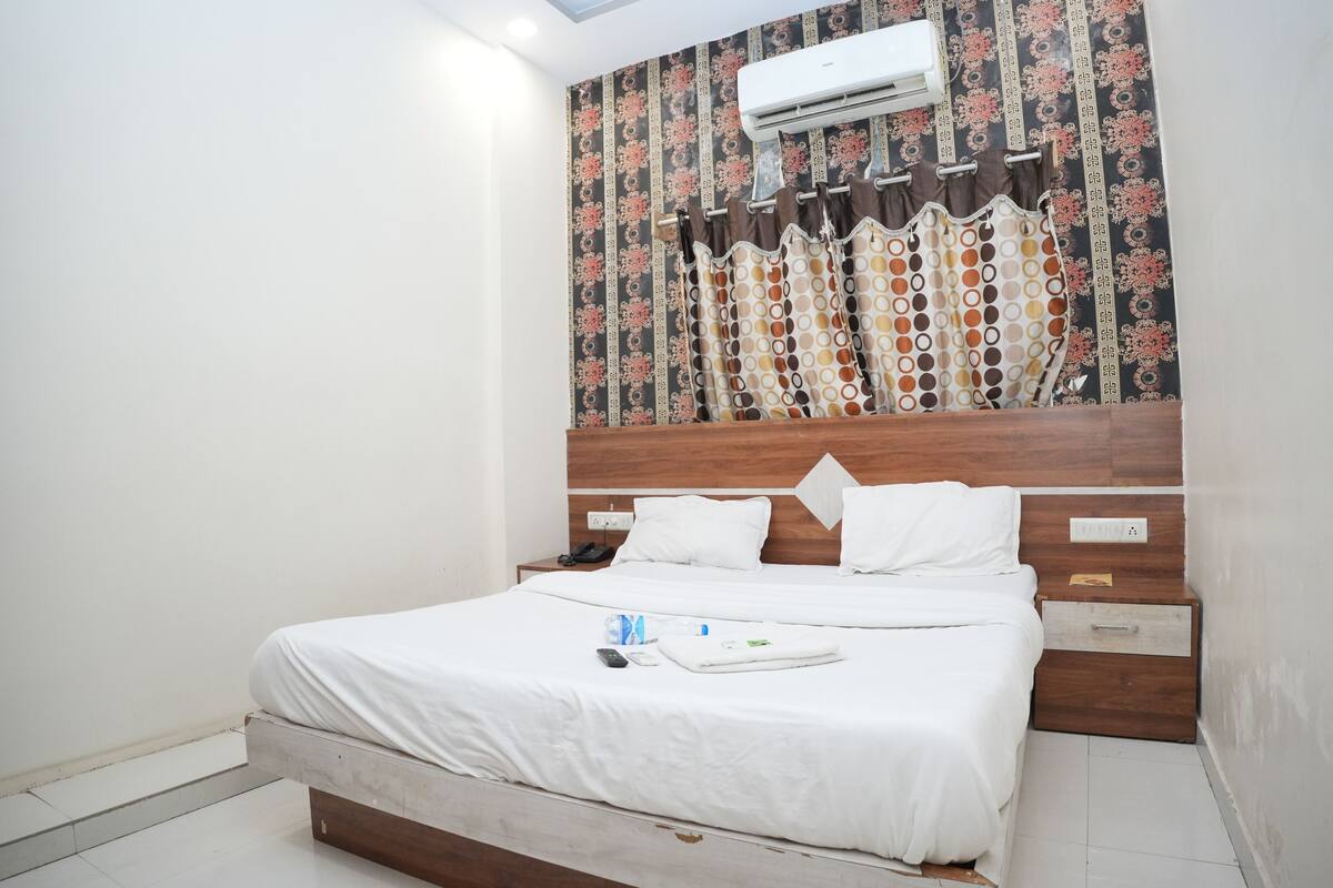 A hotel room with a bed, a wooden headboard, an air conditioner, patterned curtains, and a small bedside table with a phone and a bottle.