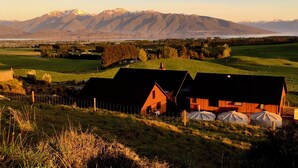 Paisatge rural amb una casa i una granja, envoltat de camps i muntanyes al fons.