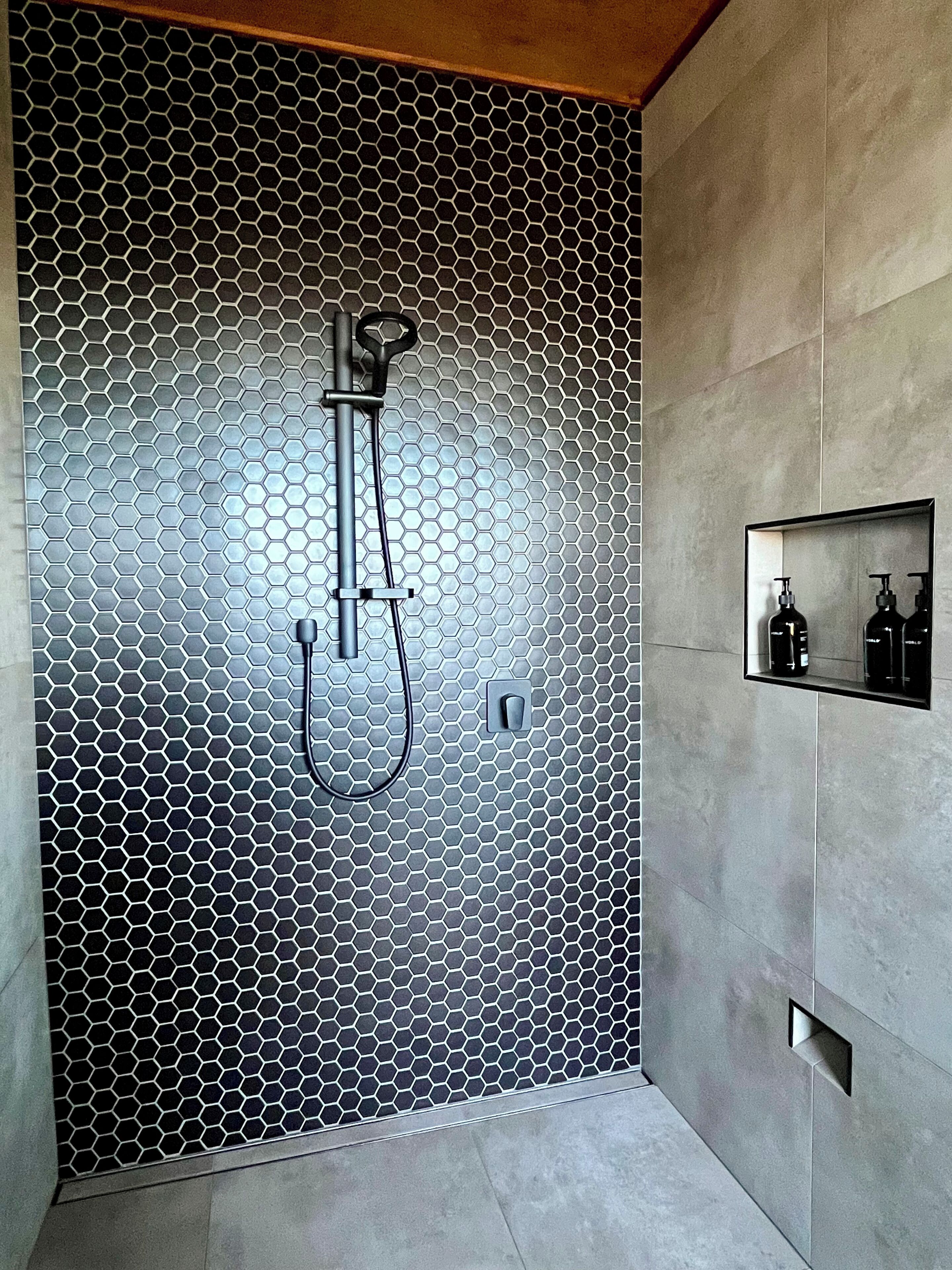 Un baño moderno con pared de azulejos hexagonales, ducha con cabezal de mano y estante empotrado con artículos de aseo.