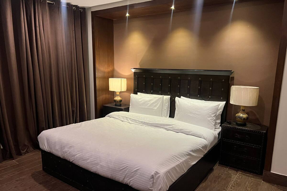 Une chambre d’hôtel avec un grand lit, une tête de lit en bois foncé, deux tables de chevet avec des lampes et un rideau sur le côté gauche.
