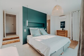 A modern bedroom with a bed, a wooden dresser, a woven lamp, and a door leading to another room.
