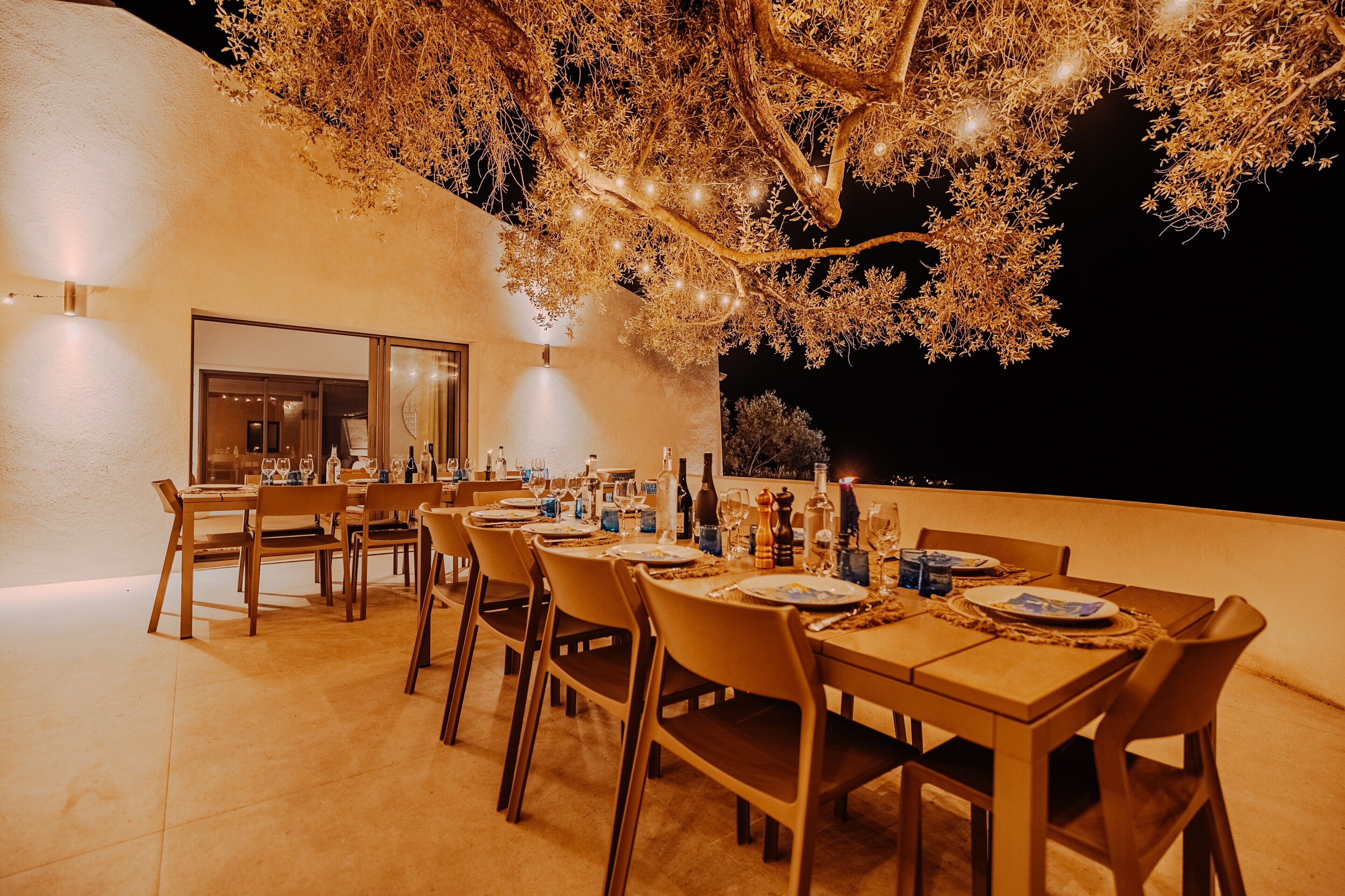 An outdoor dining area with tables set for a meal, surrounded by chairs, and a large tree overhead.
