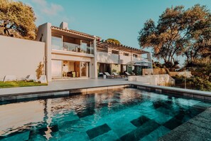 A modern two-story house with a swimming pool, outdoor seating, and a view of trees.