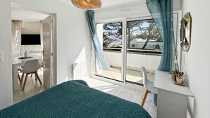A bedroom with a bed, a desk, a chair, and a balcony with a view. - Ploemeur T2 with Balcony, Garage 30m to Beach (Ploemeur)