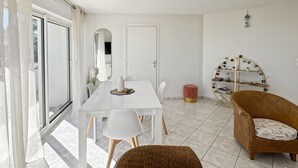 A room with a white dining table, chairs, a wicker armchair, a small red ottoman, a white door, and a wall-mounted shelf with decorative items. - Ploemeur T2 with Balcony, Garage 30m to Beach (Ploemeur)