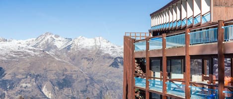 A multi-story building with balconies, set against a backdrop of snow-capped mountains.