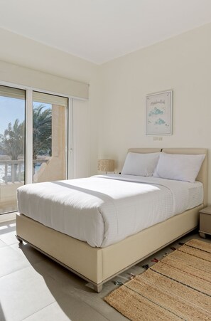 A bedroom with a bed, a nightstand, a lamp, a framed artwork, and a view of palm trees through a sliding door. - Vesta - Luxury Villa - 4BR - Joubal (El Gouna)