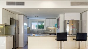 A modern kitchen with white cabinetry, stainless steel appliances, granite countertops, and two black bar stools. - Vesta - Luxury Villa - 4BR - Joubal II (El Gouna)