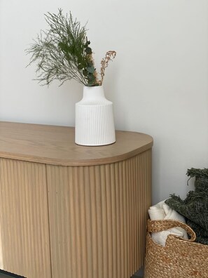 Un vase blanc à côtes, orné de feuillage vert, est posé sur un meuble en bois devant un mur uni.