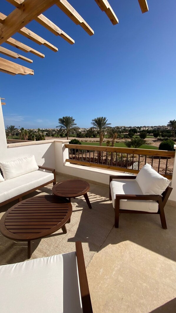Un espace extérieur aménagé, baigné de lumière, avec un canapé blanc, deux tables en bois et deux chaises, offrant une vue sur un jardin paysager et des bâtiments situés au loin.