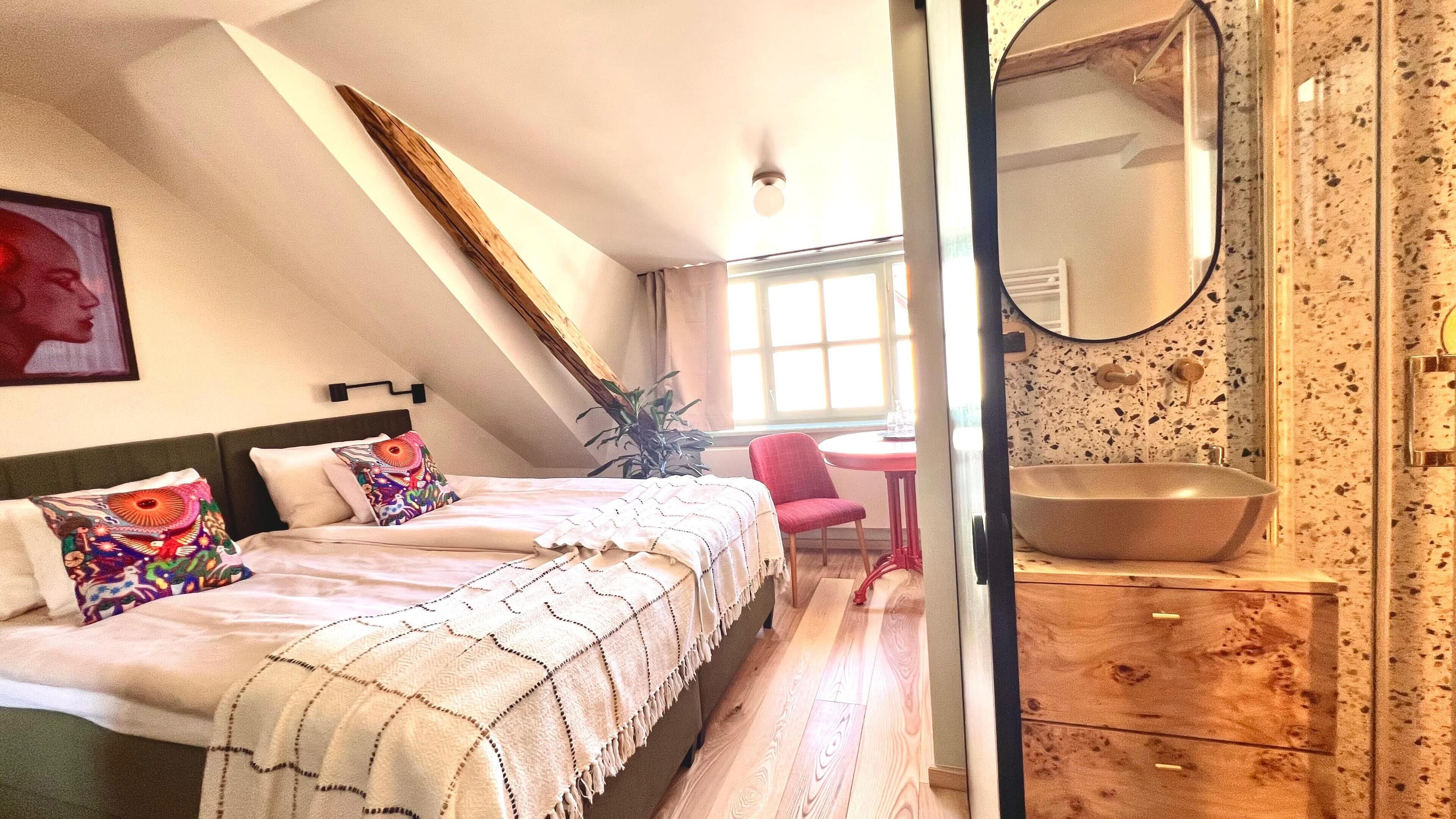 A bedroom with a bed, a desk, a chair, a plant, and a bathroom with a sink and mirror.