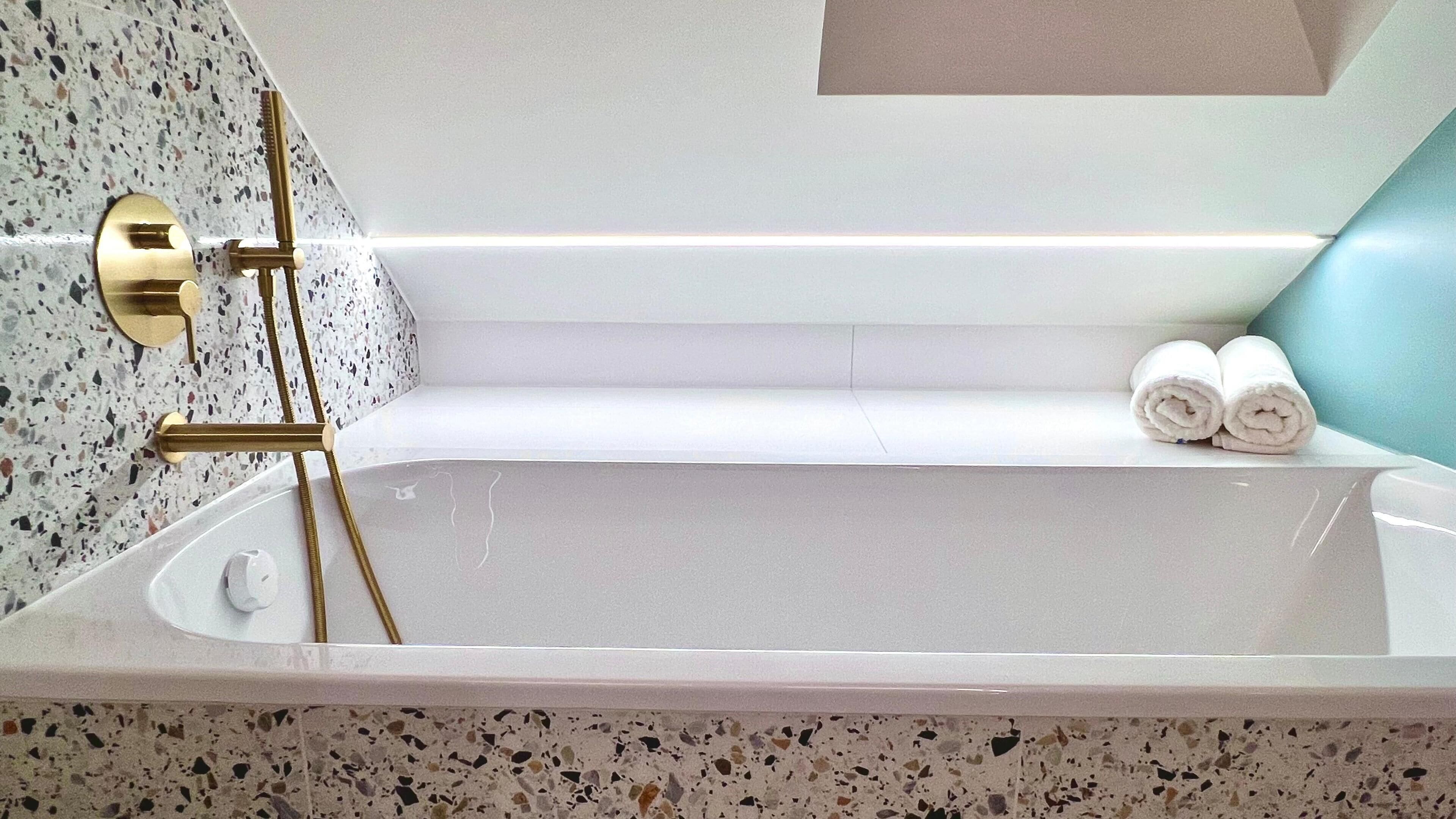 Eine Wanne mit goldenem Wasserhahn, einem Handtuchhalter mit Handtüchern und einer Wand mit geflecktem Fliesenbelag.