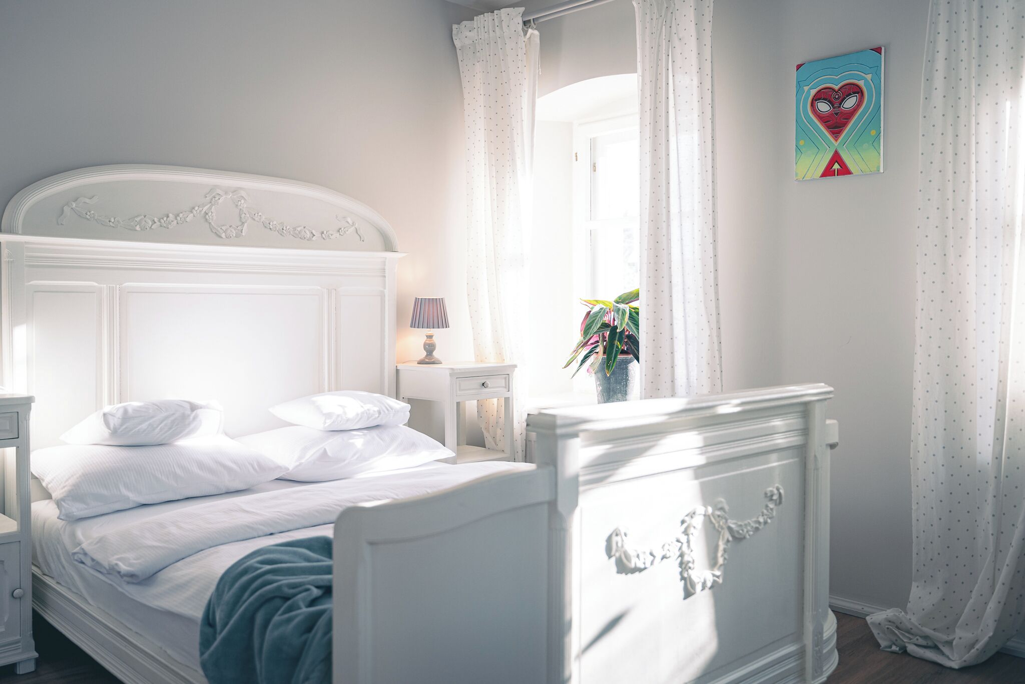 A bedroom with a white bed, a nightstand, a lamp, a window with curtains, and a wall decoration.