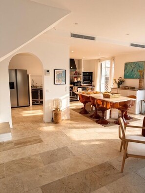 A spacious dining area with a large wooden table and chairs, a kitchen in the background, and a staircase leading to another level.