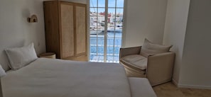 A bedroom with a large bed, a chair, a wardrobe, and a view of the marina through a window.