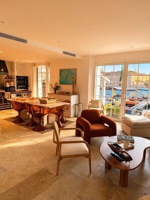 Uma sala de estar ampla com mesa de jantar, assentos confortáveis e grandes janelas com vista para um porto de iates.