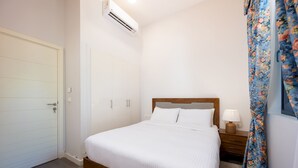 A bedroom with a bed, a nightstand, a lamp, a floral curtain, and an air conditioning unit. - Vesta - Luxury Villa - 4BR - Cyan (El Gouna)
