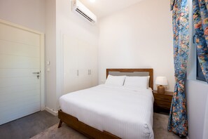 A bedroom with a bed, a nightstand, a lamp, a floral curtain, and an air conditioning unit. - Vesta - Luxury Villa - 4BR - Cyan (El Gouna)