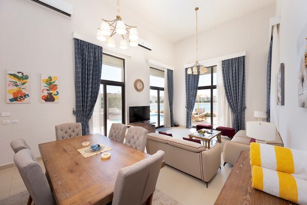 A modern living room with a wooden dining table, beige sofas, and a TV. - Vesta - Luxury Villa - 4BR - Cyan (El Gouna)