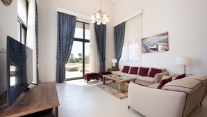 A modern living room with a flat-screen TV, a wooden coffee table, a beige sofa, and a red armchair. - Vesta - Luxury Villa - 4BR - Cyan (El Gouna)