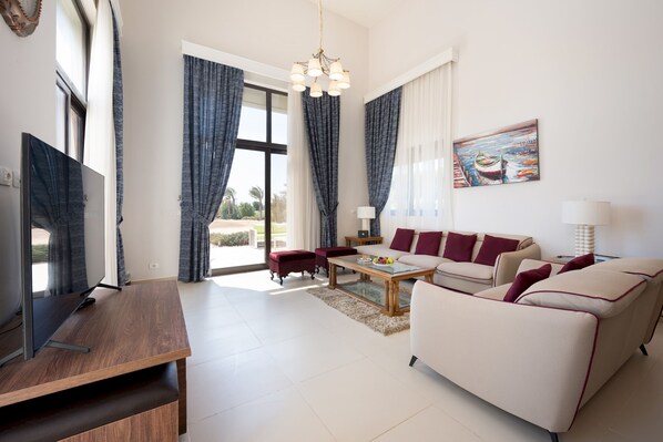 A modern living room with a flat-screen TV, a wooden coffee table, a beige sofa, and a red armchair. - Vesta - Luxury Villa - 4BR - Cyan (El Gouna)