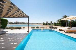 A swimming pool with lounge chairs and umbrellas, a pergola, and a view of the beach and buildings in the background. - Vesta - Luxury Villa - 4BR - Cyan (El Gouna)