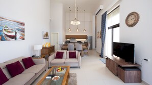 A modern living room with a sofa, coffee table, and TV. A dining area with chairs and a table is visible in the background. - Vesta - Luxury Villa - 4BR - Cyan (El Gouna)