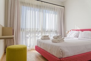 A neatly made bed with white linens and red accents, a small green ottoman, and sheer curtains allowing natural light. - Vesta - Luxury APT - 2BR - Kamaran (El Gouna)