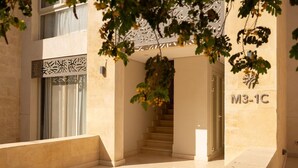 A building entrance with a staircase leading to the upper floor, a decorative wall with a sign, and a tree in the foreground. - Vesta - Luxury APT - 2BR - Mangrovy (El Gouna)