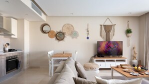 A modern living room with a beige sofa, a wooden dining table, and a flat-screen TV. There is a kitchen area with a stove and cabinets to the left. - Vesta - Luxury APT - 2BR - Mangrovy (El Gouna)