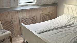 Quarto com teto inclinado, painéis de madeira, claraboia, cama com roupa de cama branca e uma cadeira.