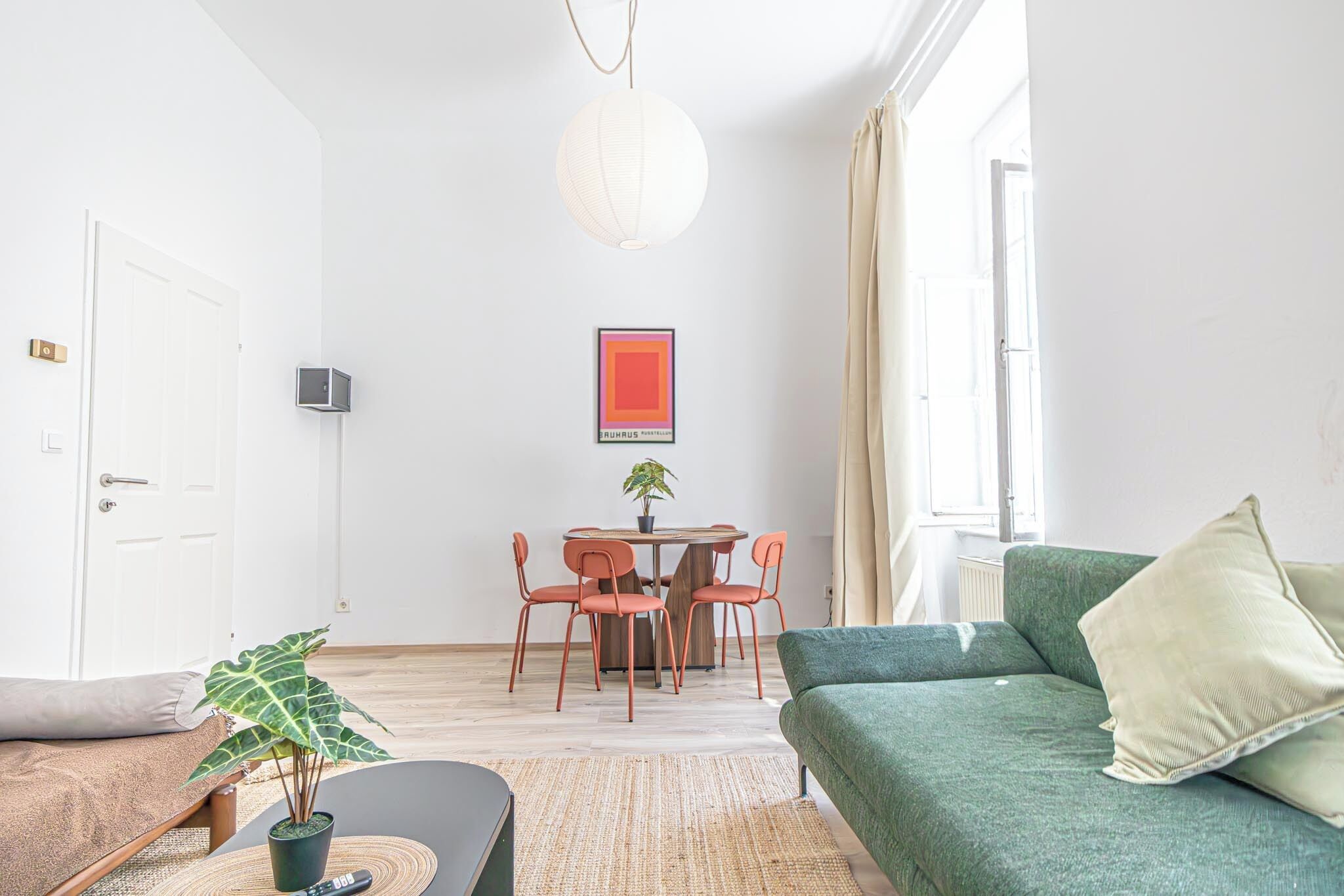 A modern living room with a green sofa, a dining table with chairs, a potted plant, and a framed artwork on the wall.