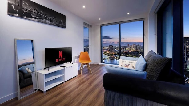 A modern living room with a large TV, a sofa, and a chair, with a city view through large windows. - Quayside Penthouse w/ City Views & Fast WiFi (Greater Manchester)