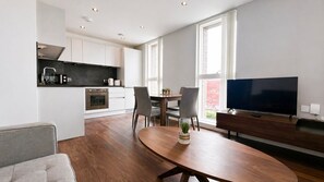 Una cocina moderna con armarios blancos, una zona de comedor con mesa y sillas de madera, y un televisor de pantalla plana sobre un mueble de madera.