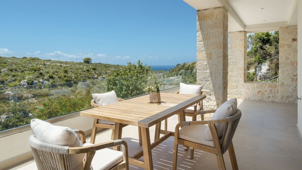 A balcony with a wooden table and chairs, overlooking a scenic landscape with greenery and a view of the sea. - Le Mont Blanc (Kounoupidiana)