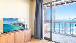 Een kamer met een televisie op een houten kast, een schuifdeur naar een balkon met uitzicht op de oceaan.