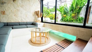 A modern living room with a glass-top table, a sofa, and a view of a lush garden through large windows. - Solla Sol Villa Canggu (Kerobokan)