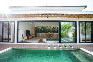 Ein modernes Haus mit einer großen Schiebeglastür, die zu einem Wohnbereich mit Küche, Pool und einer Holzterrasse führt.