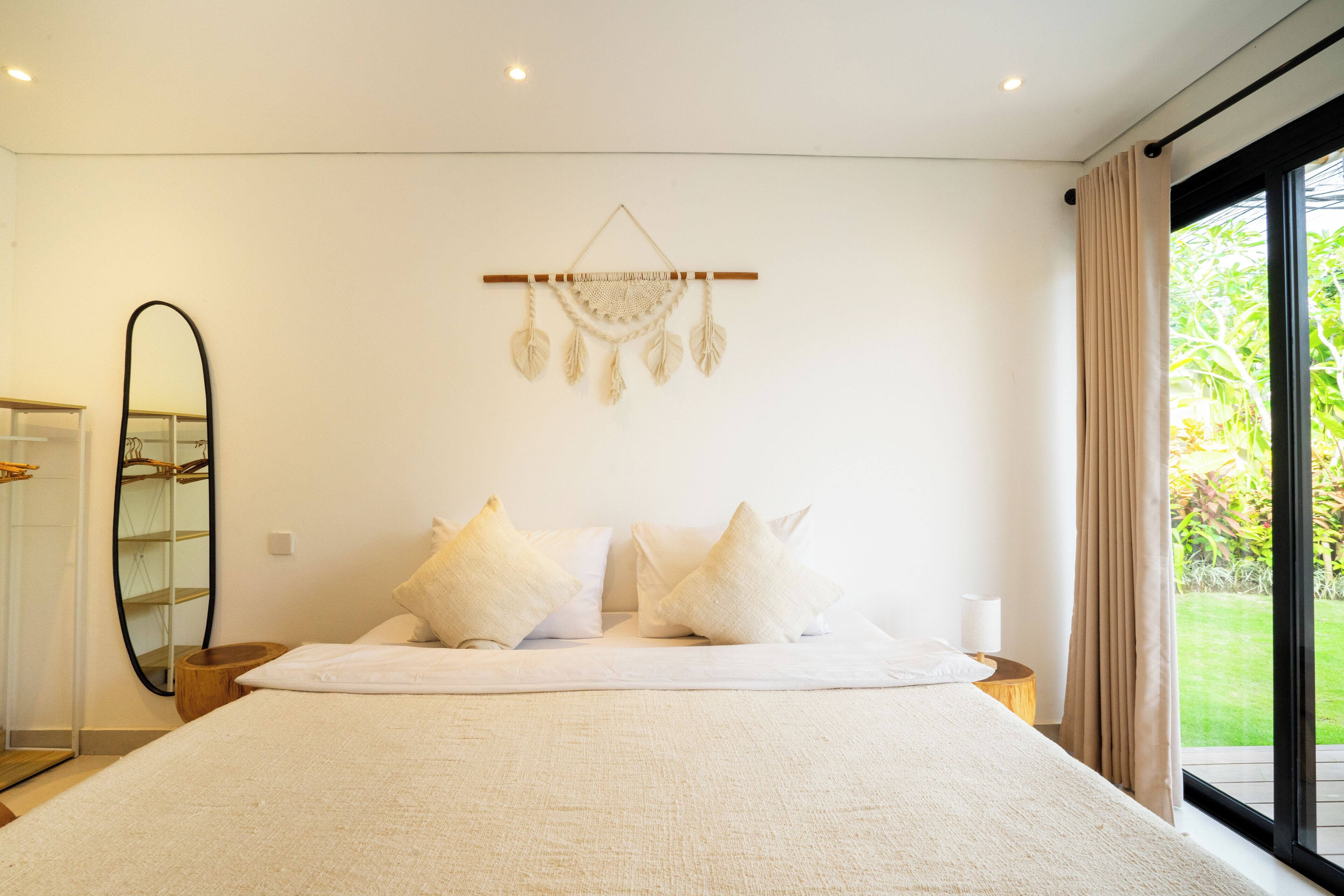 Un dormitorio con una cama grande, una mesita de noche, un adorno de macramé en la pared, un espejo y vistas a la vegetación a través de una ventana.