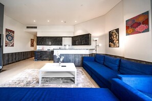 A modern living room with a blue sofa, a white coffee table, and a kitchen area with dark cabinets and a marble backsplash. - Vesta - Luxury APT - 1BR - Porto Cairo (New Cairo)