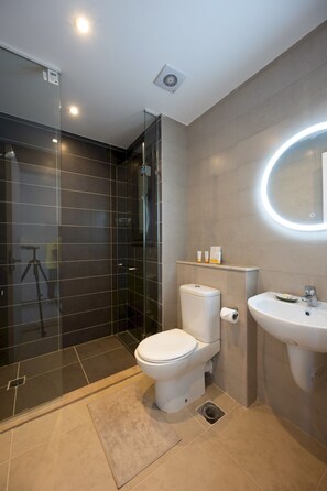 A modern bathroom with a glass shower enclosure, a toilet, a sink, and a mirror with a circular light. - Vesta - Luxury APT - 2BR - Gcribs III (El Gouna)
