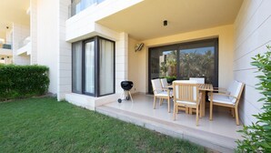A modern outdoor patio with a wooden table and chairs, a barbecue grill, and a glass door leading to a balcony. - Vesta - Luxury APT - 2BR - Gcribs III (El Gouna)
