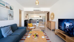 A modern living room with a blue sofa, a wooden coffee table with fruit, wicker chairs, a TV, and a kitchen in the background. - Vesta - Luxury APT - 2BR - Gcribs III (El Gouna)