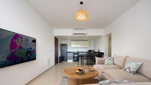 Ein modernes Wohnzimmer mit großem Flachbildfernseher, einem Holztisch, einem beigefarbenen Sofa und einer Küche im Hintergrund.